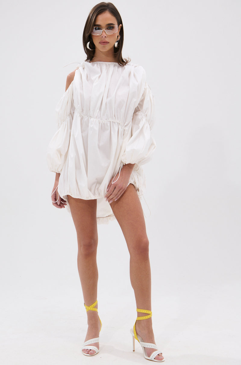 A woman wearing the INSTA SAW IT FIRST ADJUSTABLE MINI DRESS—off-white, summer-ready, with puffed sleeves and shoulder cutouts—pairs it with yellow-strapped heels and sunglasses, standing against a plain white background.