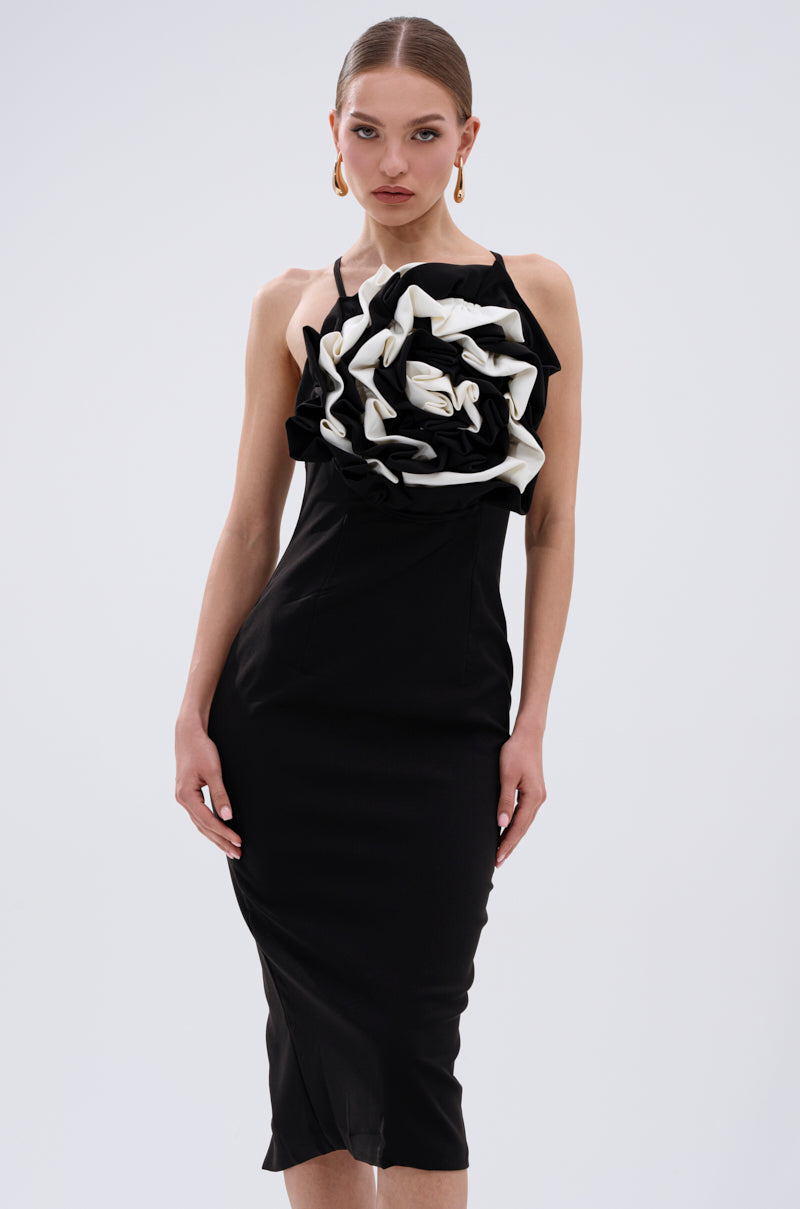 A woman wearing the MAYBE SOMEDAY MIDI DRESS WITH FLOWER APPLIQUE, a curve-hugging black midi dress with a bold white and black flower on the bodice, stands against a plain background, her hair pulled back and gold earrings visible. 0