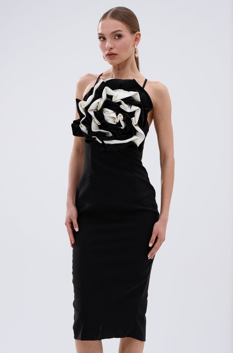 A woman stands against a plain light background wearing the MAYBE SOMEDAY MIDI DRESS WITH FLOWER APPLIQUE, a curve-hugging midi dress featuring a bold black and white sculptural flower on the bodice. 1