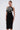 A woman stands against a plain light background wearing the MAYBE SOMEDAY MIDI DRESS WITH FLOWER APPLIQUE, a curve-hugging midi dress featuring a bold black and white sculptural flower on the bodice.