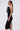 A woman in profile wears the MAYBE SOMEDAY MIDI DRESS WITH FLOWER APPLIQUE, featuring a curve-hugging fit, white and black ruffled flower detail on the chest, side slit hem, gold hoop earrings, and long brown hair in a low ponytail.