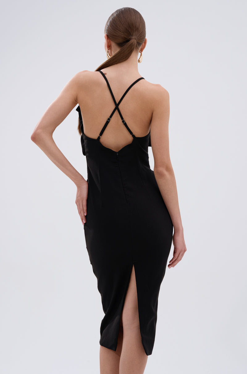 A woman with long, straight hair models the MAYBE SOMEDAY MIDI DRESS WITH FLOWER APPLIQUE—a fitted, sleeveless black midi dress with thin crossed straps and a back slit. She stands facing away, hand on her hip against a plain light background. 3
