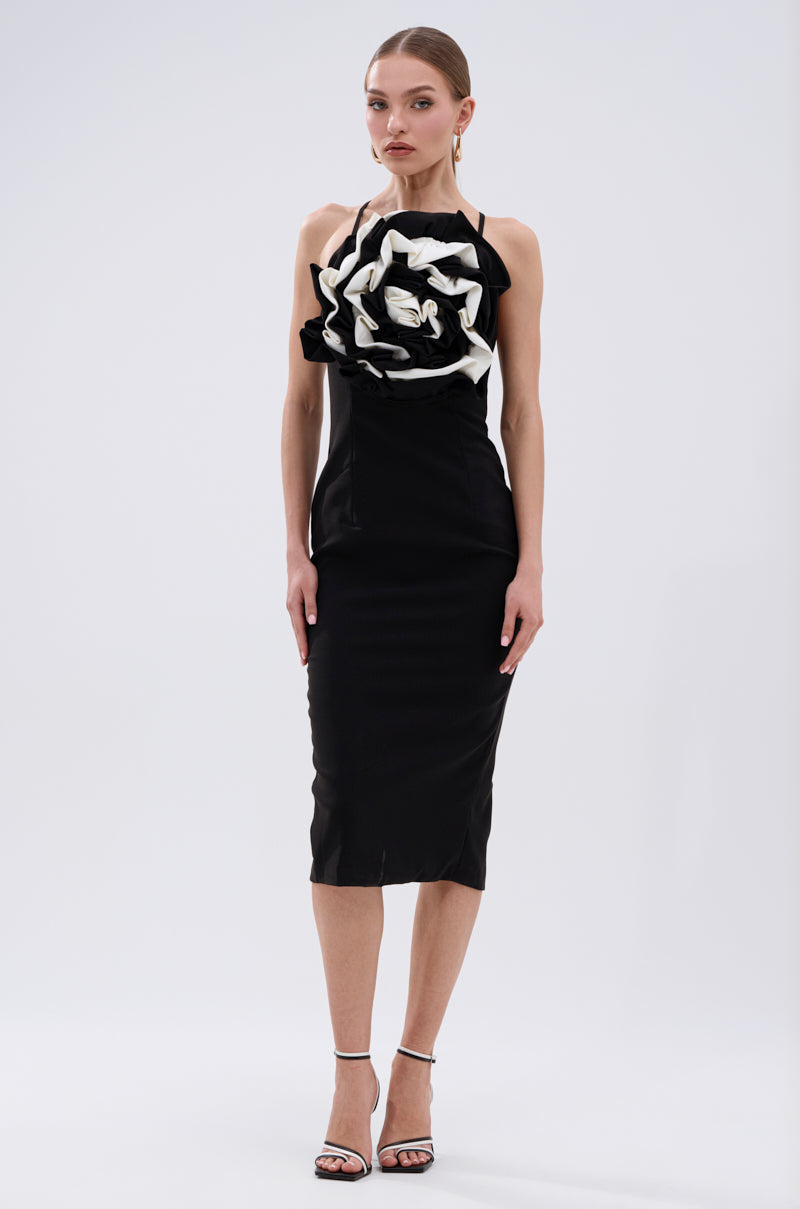 A woman wears the MAYBE SOMEDAY MIDI DRESS WITH FLOWER APPLIQUE— a curve-hugging, sleeveless black midi dress with a bold black-and-white flower detail—paired with heels against a plain white background. 5