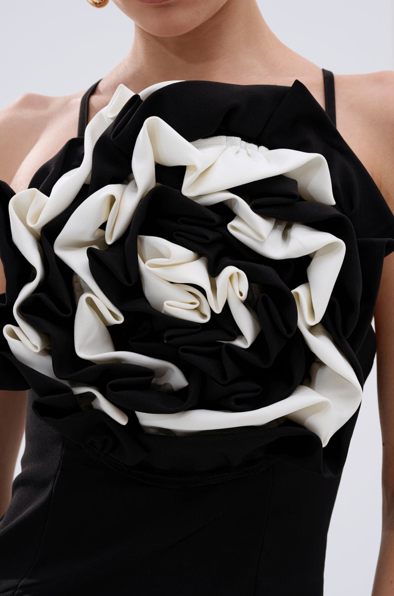 A close-up of a woman wearing the MAYBE SOMEDAY MIDI DRESS WITH FLOWER APPLIQUE, a curve-hugging black midi dress detailed with a dramatic, sculpted flower and layered ruffled petals on the chest; her face is partially out of frame. 6