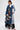 A woman models the MILA PATCH DENIM TRENCH—a long denim coat with embroidered patches and letters—over a white blouse and black lace skirt, styled with red shoes and glasses against a plain white background for bold streetwear flair.