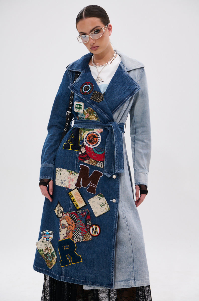 Model wears the MILA PATCH DENIM TRENCH—a long, belted denim coat with colorful embroidered patches—styled with glasses, a necklace, and tied-back hair against a plain white background for a cool streetwear vibe.