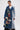 Model wears the MILA PATCH DENIM TRENCH—a long, belted denim coat with colorful embroidered patches—styled with glasses, a necklace, and tied-back hair against a plain white background for a cool streetwear vibe.