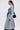 A woman in the MILA PATCH DENIM TRENCH, clear glasses, and a black purse stands in profile against a white background, showcasing modern streetwear style.