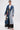 Wearing the MILA PATCH DENIM TRENCH with embroidered patches over a graphic tee, black lace skirt, red and white shoes, and clear glasses, a person stands against a plain white background showcasing bold streetwear style.