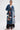A woman models the MILA PATCH DENIM TRENCH, a long denim coat with colorful patches, over a black lace skirt. She pairs it with slicked-back hair, sunglasses, and red-and-white shoes against a plain white background.