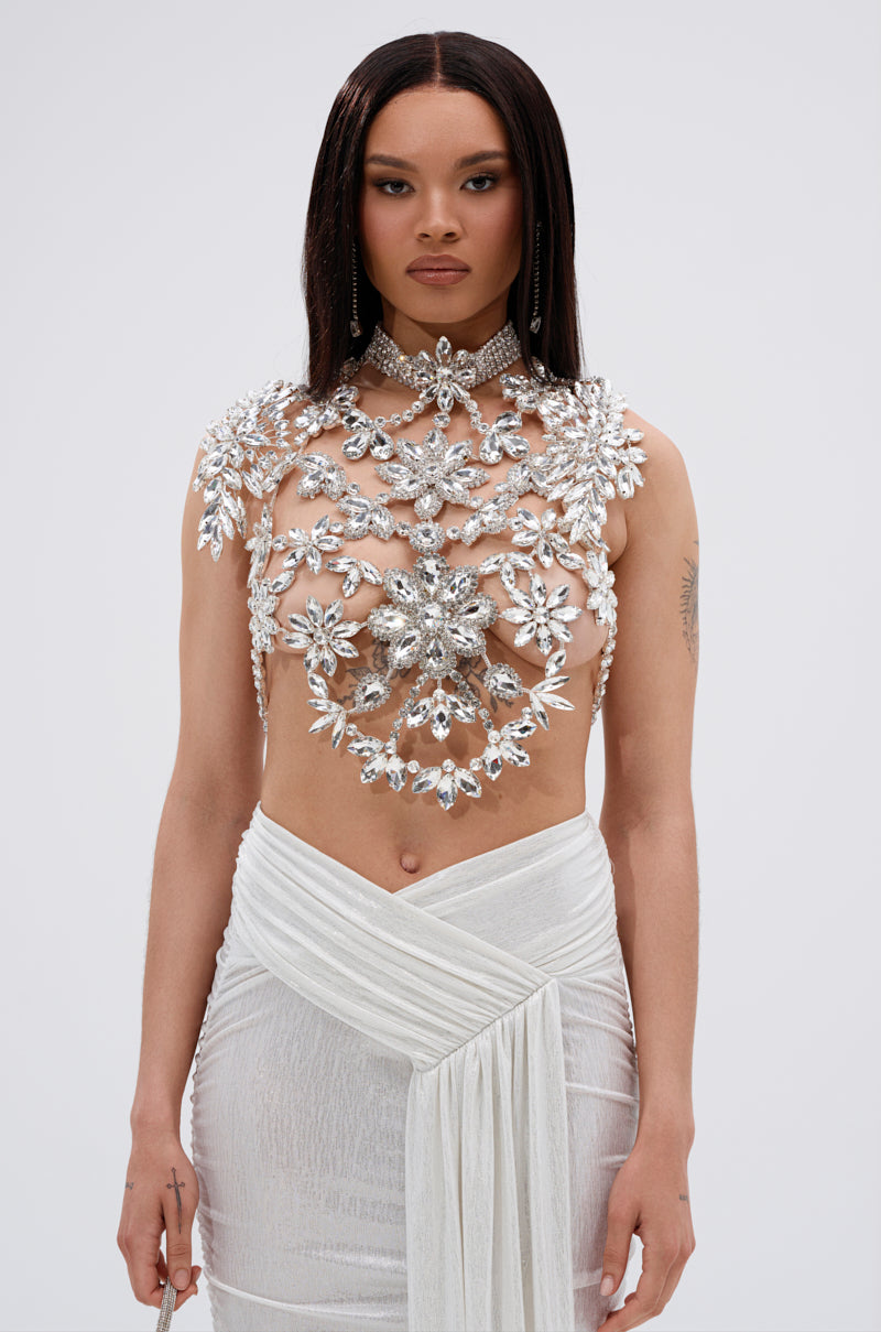 A woman with long, straight hair stands against a plain background, wearing the ICE QUEEN RHINESTONE TOP— a sheer, crystal-embellished white piece—paired with a draped, shimmery skirt. She has a neutral expression.