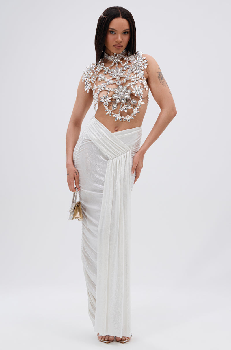 A woman poses in the ICE QUEEN RHINESTONE TOP, its crystal embellishments shimmering under the light. She pairs it with a white draped skirt and gold purse, standing elegantly against a plain white background.