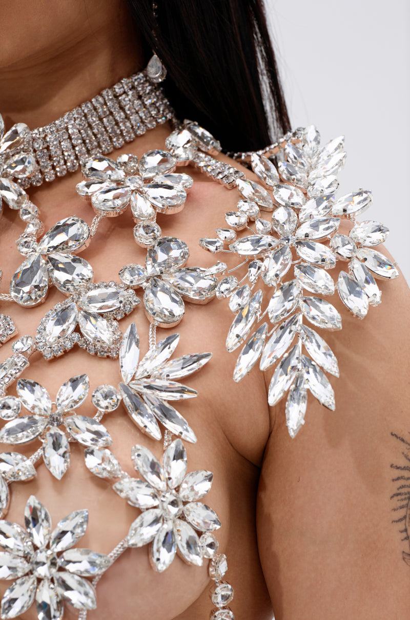 A close-up of someone wearing the ICE QUEEN RHINESTONE TOP, a harness-style piece adorned with sparkling crystal gems in floral patterns, showing some skin and part of a tattooed arm.