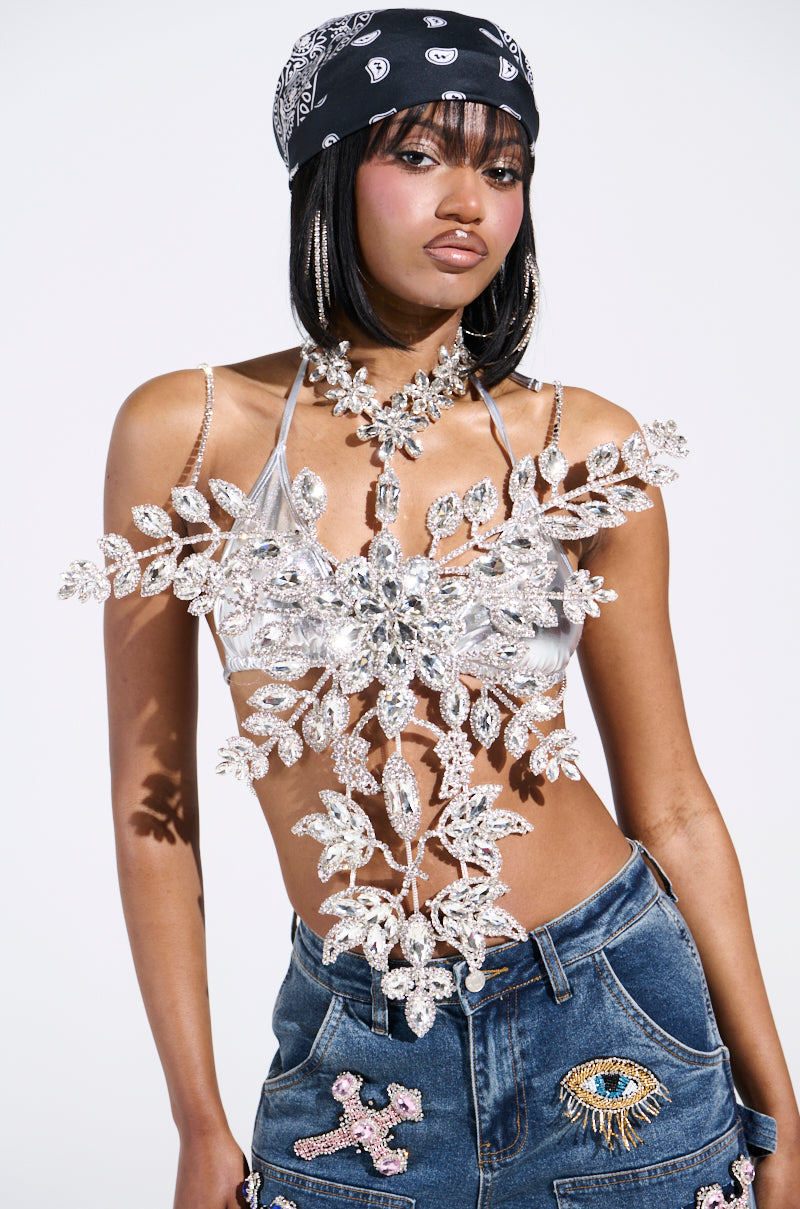 Someone dazzles in the BLINDED BY THE LIGHTS RHINESTONE TOP, paired with heavily jeweled, floral-inspired rhinestone jewelry adorning their upper body. A bandana crowns their head while decorative patch denim jeans complete the look against a plain white backdrop.