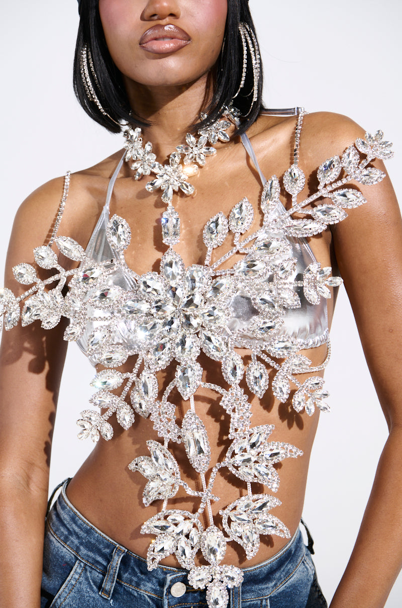 A person in the dazzling BLINDED BY THE LIGHTS RHINESTONE TOP, featuring intricate floral patterns, pairs it with denim jeans. Their long dark hair cascades beautifully against the plain white backdrop, highlighting the outfit's elaborate design.
