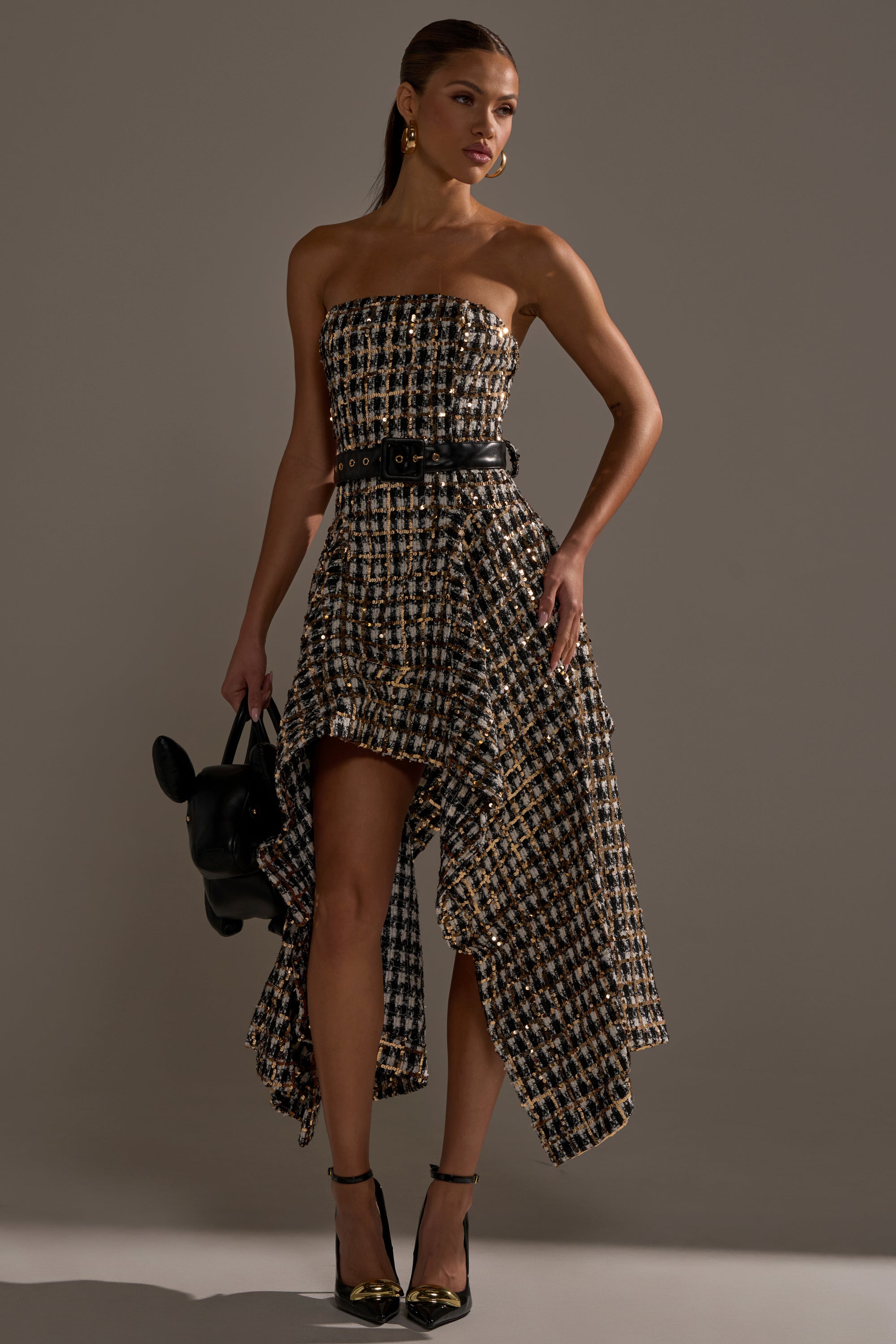 A woman models the PURE ROYALTY BROCADE HIGH LOW TWEED DRESS WITH SEQUINS, featuring a strapless, asymmetrical design and black belt, styled with an animal-shaped black bag and ankle-strap heels to highlight the chic high-low hem.