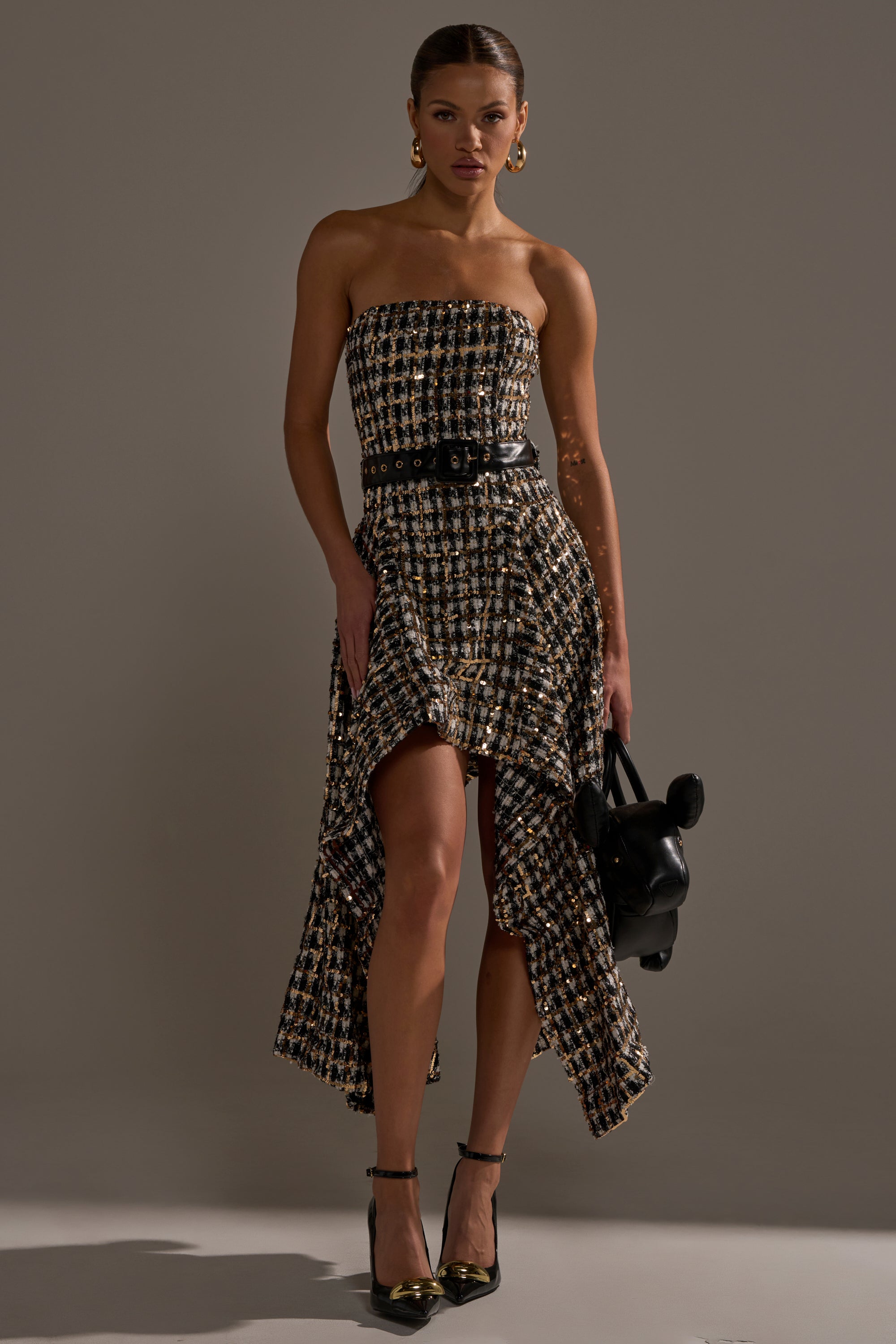 A woman models the PURE ROYALTY BROCADE HIGH LOW TWEED DRESS WITH SEQUINS—strapless, high-low, and cinched with a black belt—styled with gold heels, hoop earrings, and a black handbag against a plain gray backdrop.