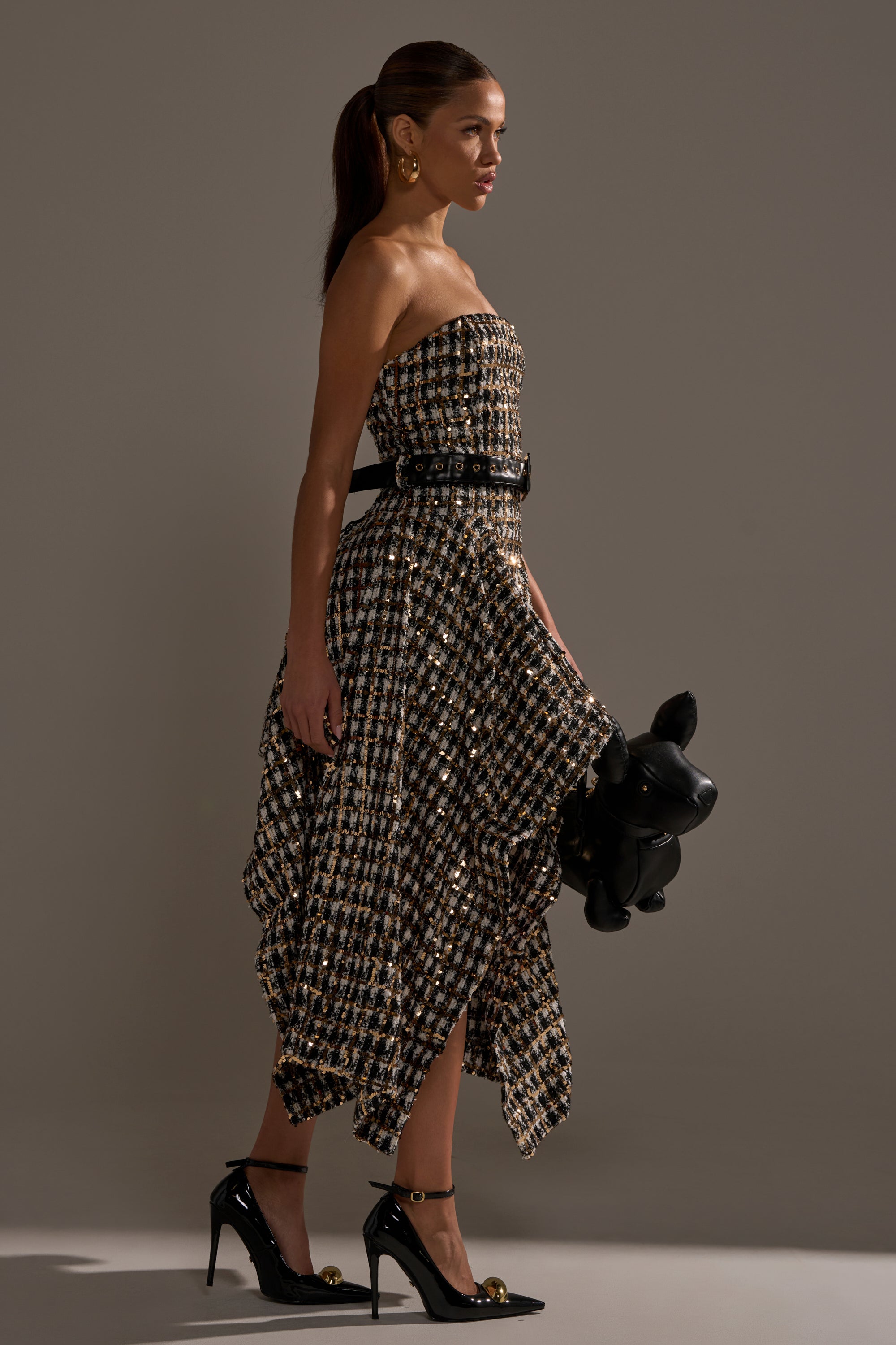 A woman in the PURE ROYALTY BROCADE HIGH LOW TWEED DRESS WITH SEQUINS, featuring a black-and-gold checkered pattern and an asymmetrical hem, stands in profile holding a black balloon dog, wearing black heels and gold hoop earrings.