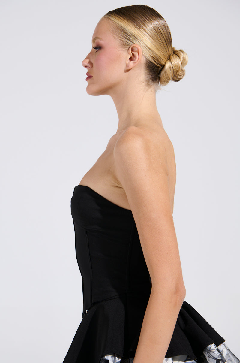 A woman with blonde hair styled in a neat bun poses in profile, highlighting the strapless design of her LADY FRIEND CORSET and silver floral skirt. The curve-hugging silhouette enhances her figure against the plain white background.