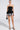 In a neutral setting, someone exudes confidence in the curve-hugging LADY FRIEND CORSET, a black strapless dress with peplum and floral-patterned hem. Strappy heels and a chic black handbag enhance the look, while styled-back hair completes this poised ensemble.