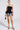 Wearing the LADY FRIEND CORSET in a strapless design, they pair it with a layered black and white ruffled skirt. A sleek black purse complements their look, along with stylish black strappy heels against a plain, light backdrop, exuding confidence.