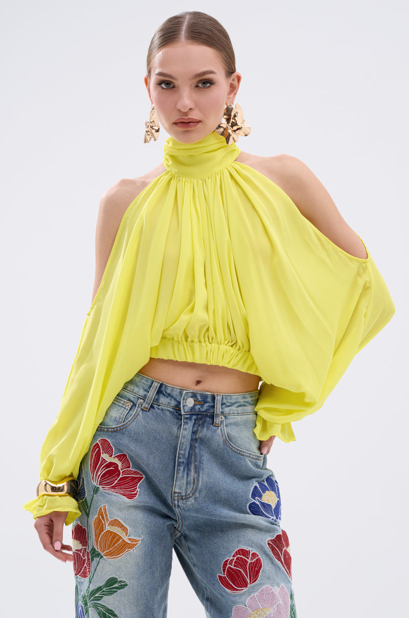 A woman models the BRIGHTEST CITY TURTLENECK BLOUSE IN YELLOW, a lightweight chiffon top with a high neck and billowy sleeves, paired with blue jeans featuring colorful floral embroidery, against a white background.