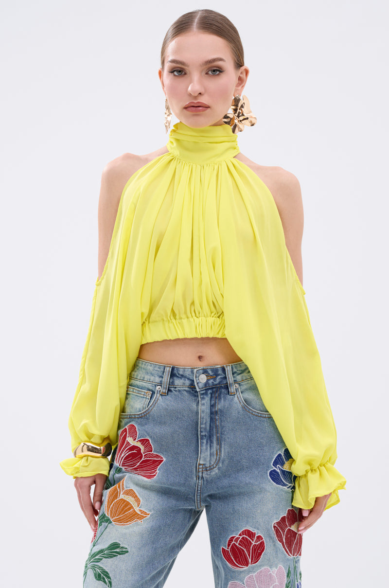 A woman wears the BRIGHTEST CITY TURTLENECK BLOUSE IN YELLOW, a lightweight chiffon piece with billowy sleeves and cropped silhouette, paired with high-waisted blue jeans with floral embroidery. She sports slicked-back hair and statement earrings.