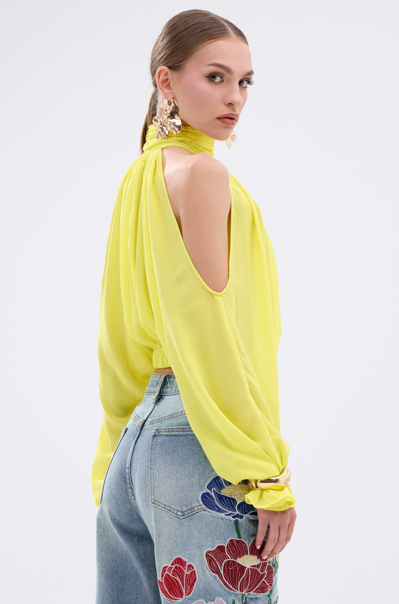 A woman models the BRIGHTEST CITY TURTLENECK BLOUSE IN YELLOW, a lightweight, open-shoulder chiffon top with a high neck, paired with light blue jeans with colorful floral embroidery on the back pocket, against a white background.