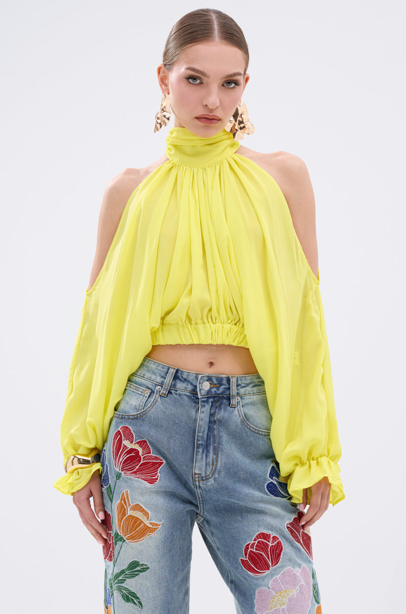 A woman wears the BRIGHTEST CITY TURTLENECK BLOUSE IN YELLOW, styled in lightweight chiffon with a vibrant hue and paired with high-waisted blue jeans featuring colorful floral embroidery.