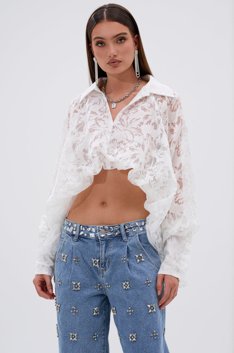 A woman wears the JACKIE RHINESTONE CROP BLOUSE, a sheer white top with a floral burnout pattern and asymmetrical high-low hem, paired with blue jeans featuring jeweled details and silver accessories.