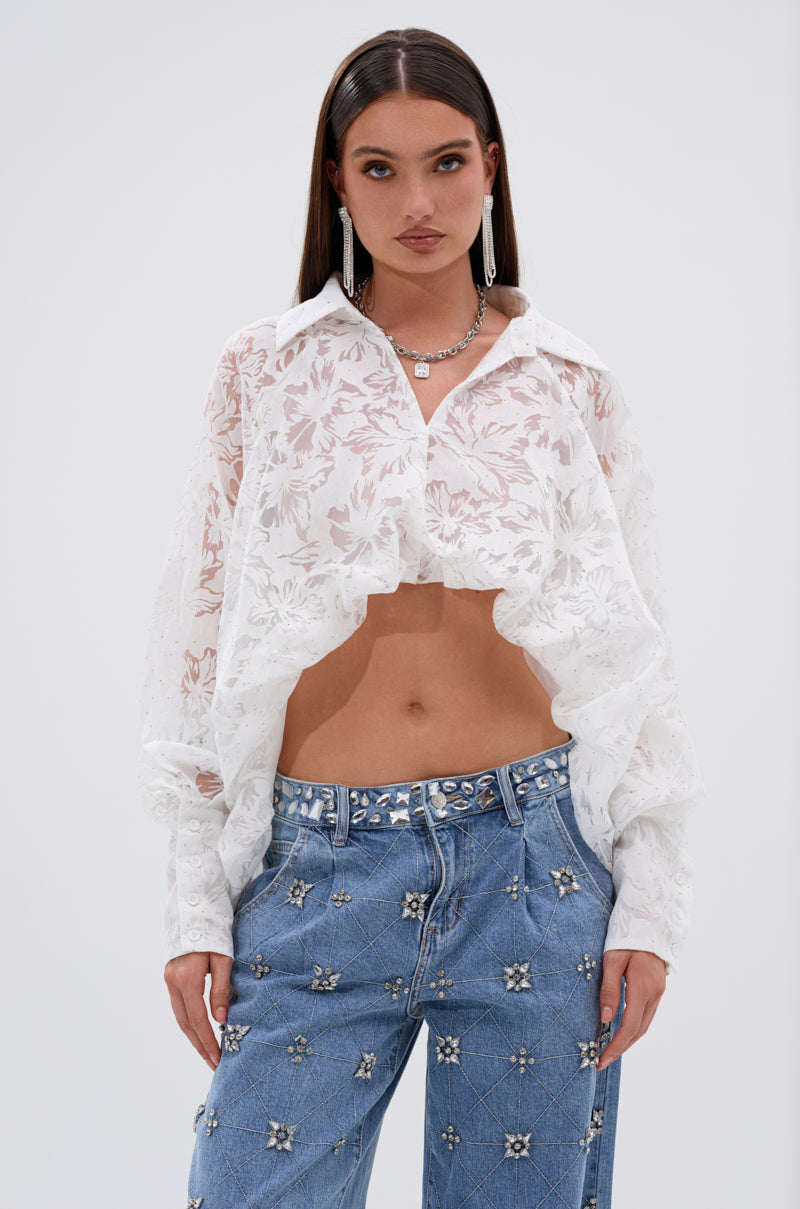 A woman with straight dark hair wears the JACKIE RHINESTONE CROP BLOUSE—a sheer white crop top with a floral burnout pattern—paired with blue jeans, long earrings, and layered necklaces against a plain background.