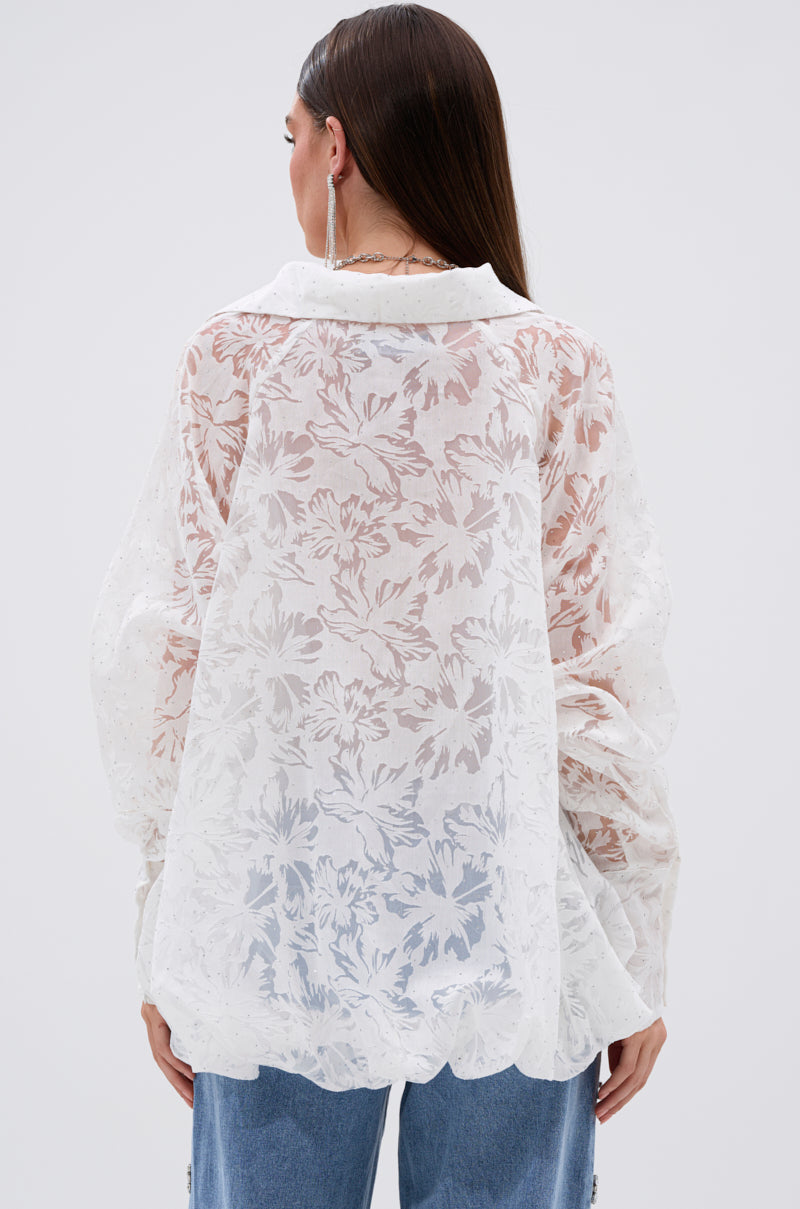 A woman with long, straight brown hair is seen from the back wearing the JACKIE RHINESTONE CROP BLOUSE—a sheer white top with floral burnout pattern and puffy sleeves—paired with light blue jeans, set against a plain light background.