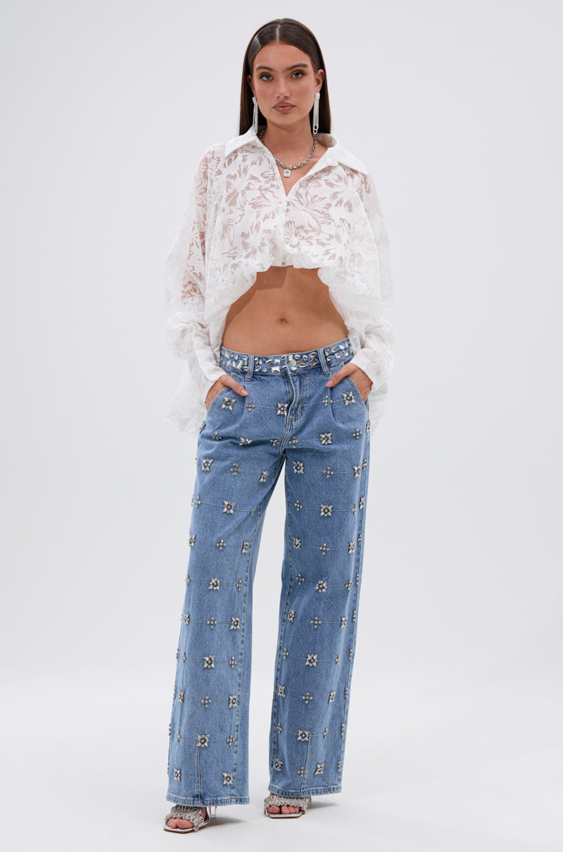 A woman poses against a plain background wearing the JACKIE RHINESTONE CROP BLOUSE, a sheer long-sleeve white lace top with floral burnout and rhinestones, high-rise light blue embellished jeans, and heeled sandals, hands in her pockets.