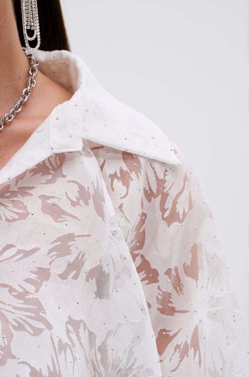 Close-up of a person wearing the JACKIE RHINESTONE CROP BLOUSE, a sheer white top with floral burnout pattern and sparkling rhinestone details, paired with a chunky chain necklace and long rhinestone earring. Plain light background.