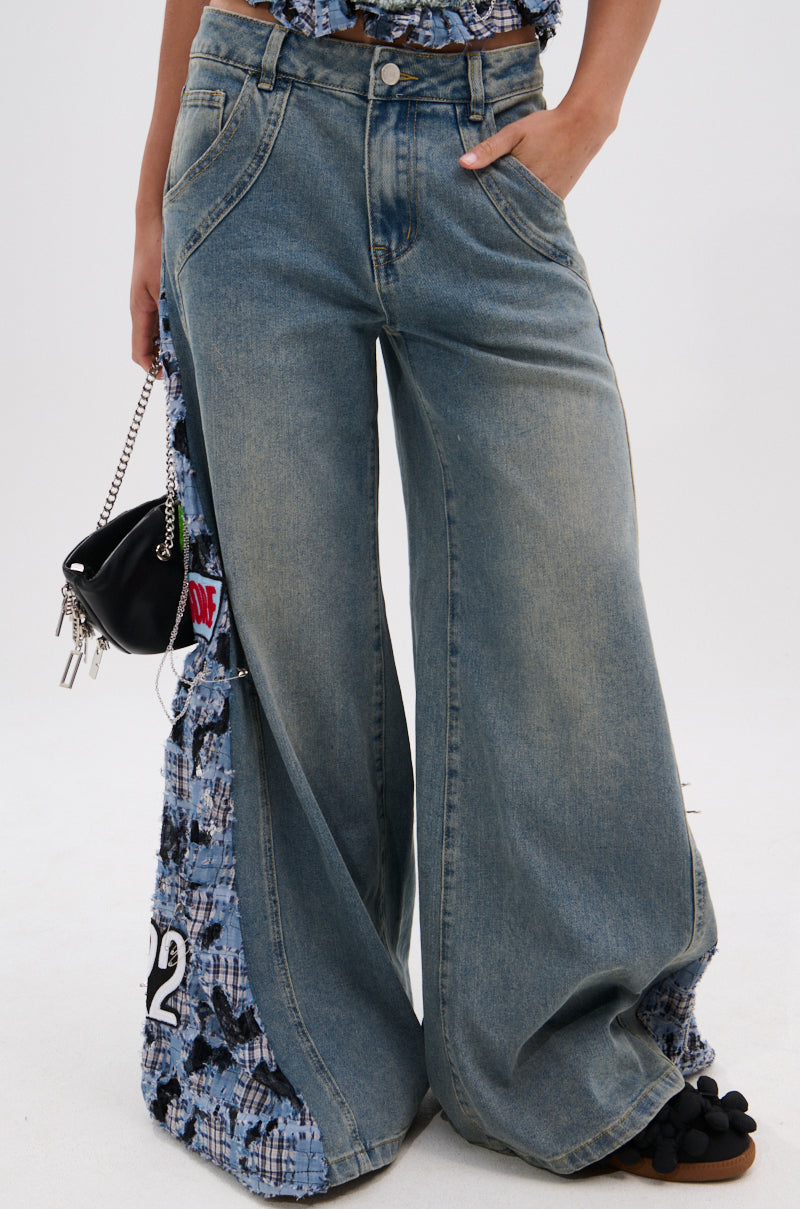 A person styled in the ON POINT PLAID EMBELLISHED DENIM PANT featuring a high-rise fit, wide leg silhouette, and plaid panel patches on the sides, paired with a patterned top and carrying a small black handbag.
