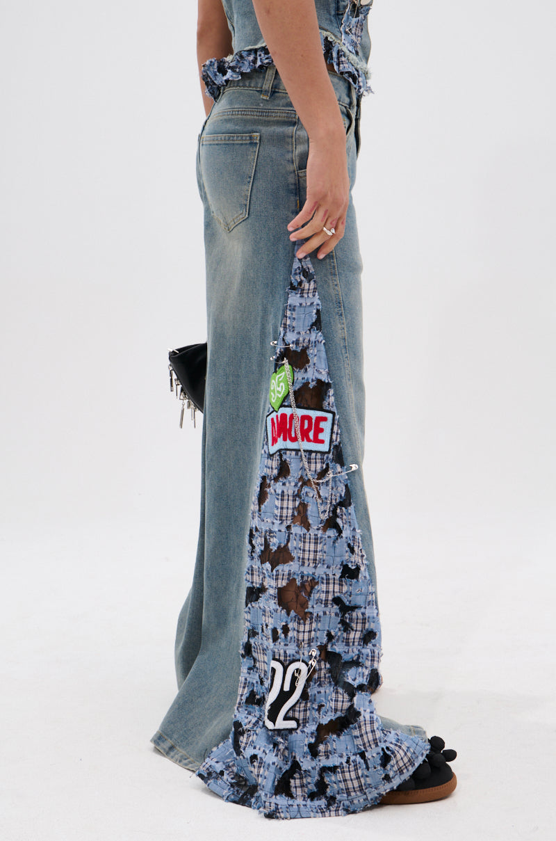 A person stands sideways in ON POINT PLAID EMBELLISHED DENIM PANT featuring wide legs, frayed lace inserts, patches, and a "22" near the hem. They hold a small black bag and wear black slide sandals against a plain white background.