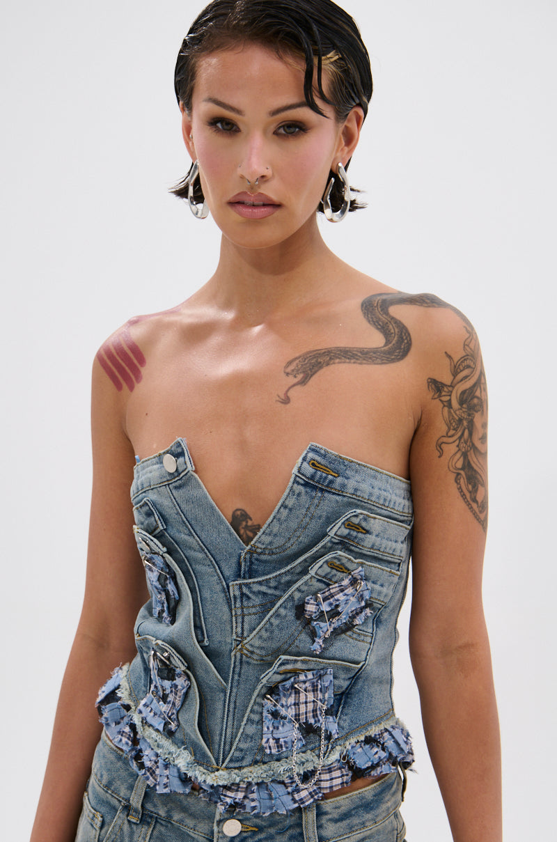 A person with slicked-back hair and visible tattoos wears the ON POINT PLAID EMBELLISHED CORSET with frayed details and matching jeans, standing against a plain light background.