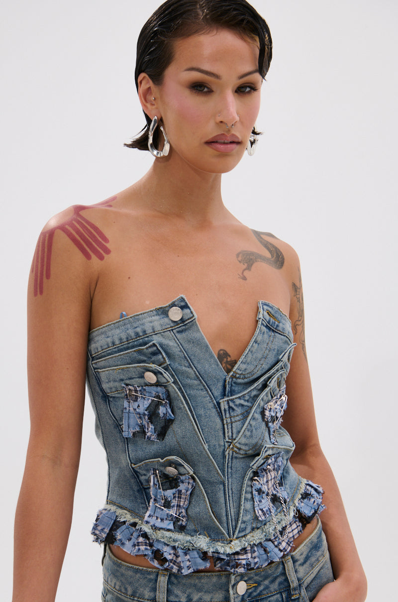 A person with short, wet-look hair and hoop earrings wears the ON POINT PLAID EMBELLISHED CORSET, featuring a strapless design with unique details. They have visible tattoos and stand against a plain light background.