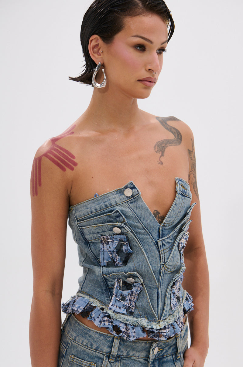 A woman with short, slicked-back hair and tattoos on her chest, arm, and shoulder wears large silver hoop earrings and the ON POINT PLAID EMBELLISHED CORSET—a strapless denim top with frayed edges and plaid patches.