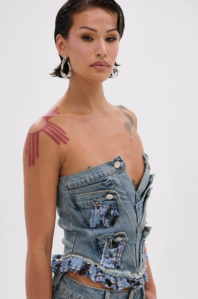 A person with short dark hair wears the ON POINT PLAID EMBELLISHED CORSET, featuring button details and ruffled edges. They accessorize with large hoop earrings, pink eyeshadow, and a red handprint tattoo on their shoulder against a plain white background.