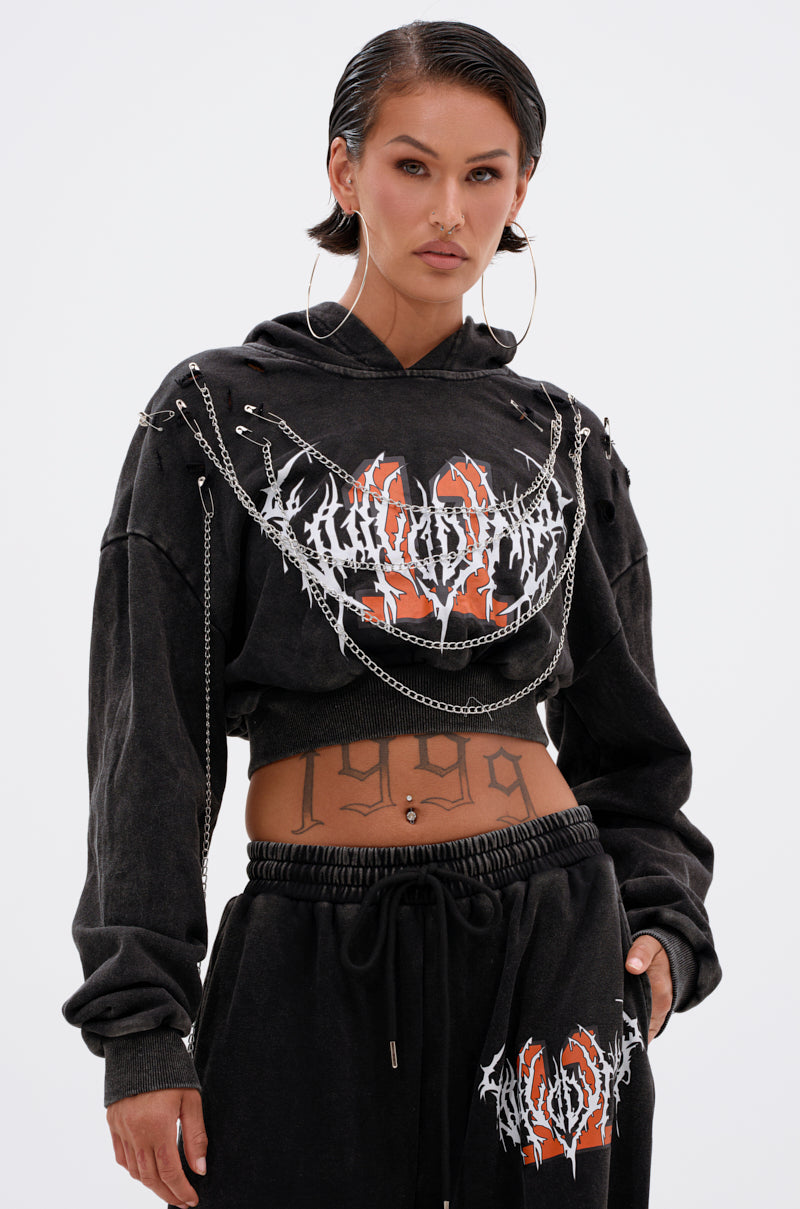 A person with slicked-back hair wears the LUCKY ELEVEN CHAIN CROP SWEATSHIRT and matching pants, featuring bold graphic prints, metal chains, large hoops, and a visible "1999" stomach tattoo. 1