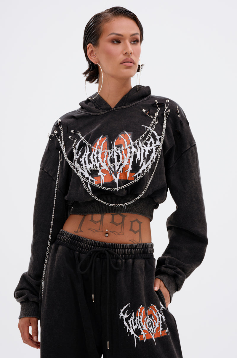 A woman stands confidently against a white background in the LUCKY ELEVEN CHAIN CROP SWEATSHIRT featuring bold white and orange graphics, metal chains, and paired with matching sweatpants and hoop earrings. 2