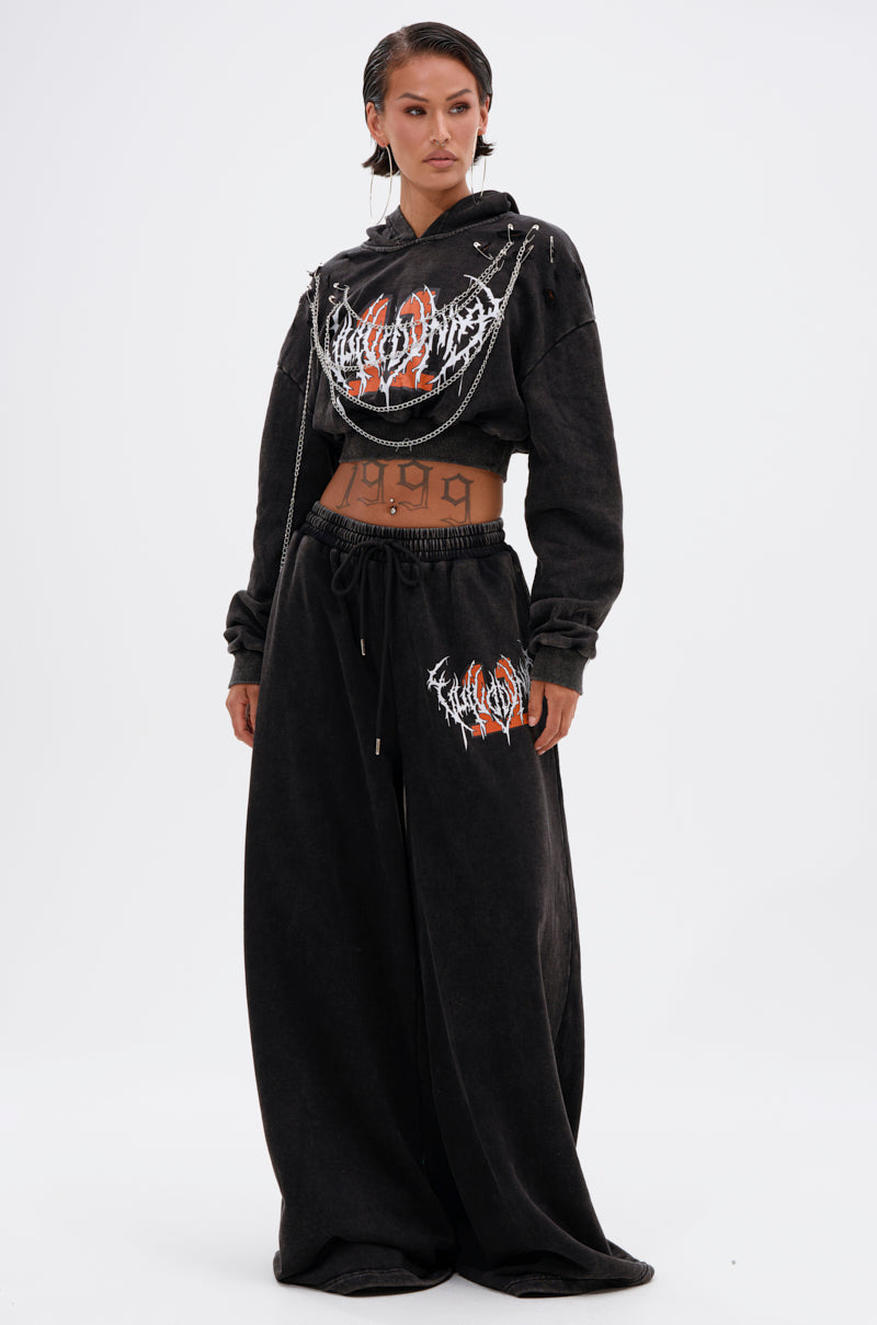 Wearing slicked-back hair, they style the LUCKY ELEVEN CHAIN CROP SWEATSHIRT with bold graphics and wide-leg black pants. A chain accessory drapes over the hoodie, revealing their midriff and a "1999" tattoo for standout streetwear flair. 5