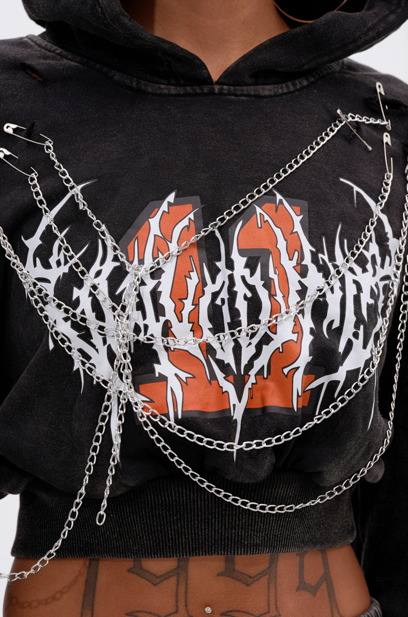 Wearing the LUCKY ELEVEN CHAIN CROP SWEATSHIRT in black with an orange and white graphic, styled with layered silver chains and safety pins across the chest, this look also highlights a tattooed midriff and a belly button piercing. 7