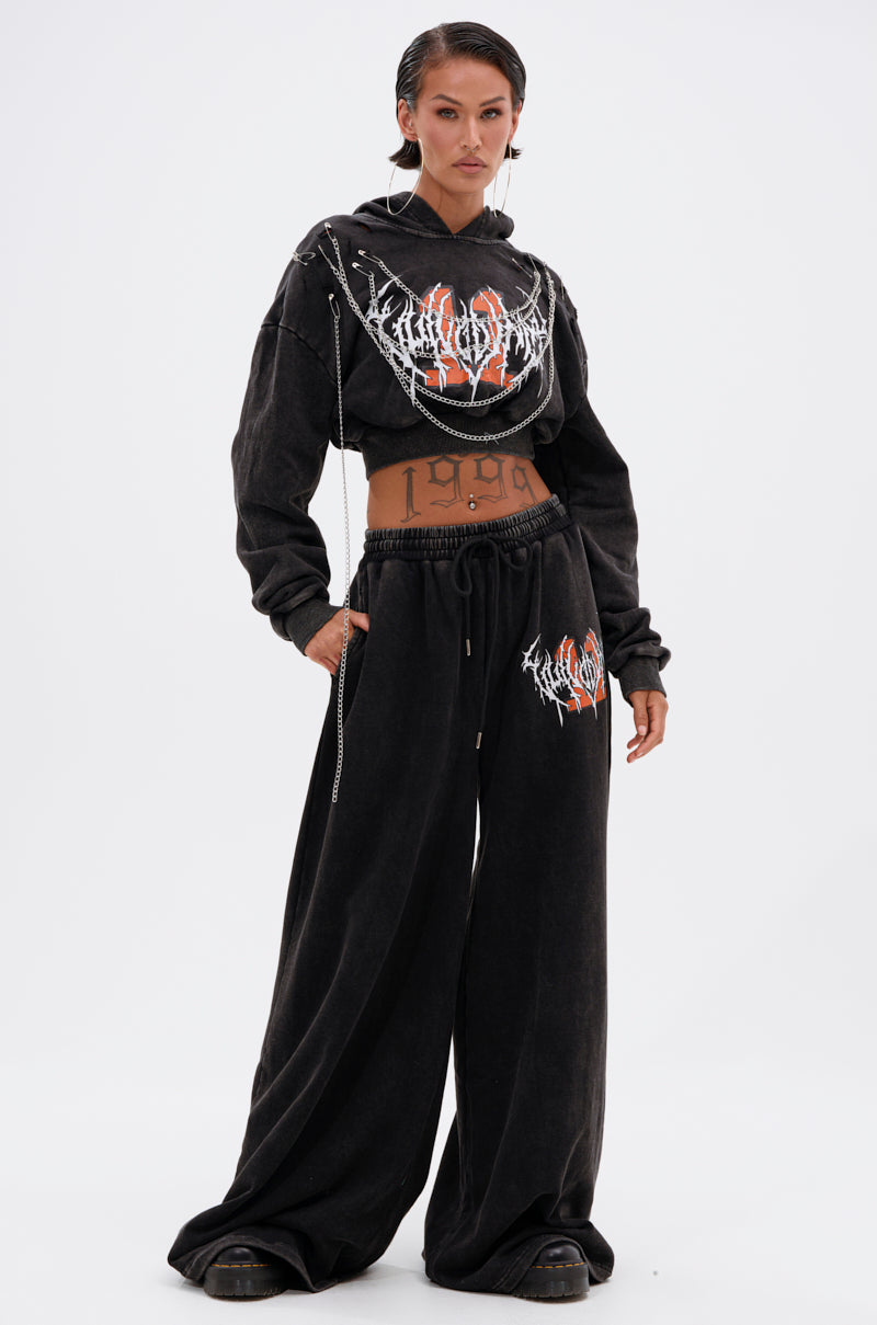 Wearing the LUCKY ELEVEN WIDE LEG JOGGER, this person stands confidently in a black cropped hoodie with white and red graphics. Slicked-back hair and "1999" on their midriff complete the edgy streetwear vibe. 0