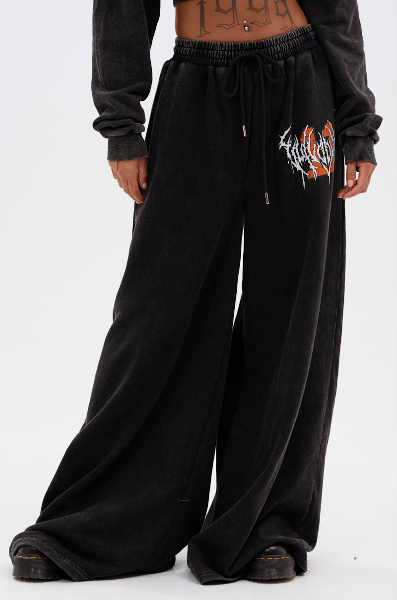 The lower half of a person is shown wearing LUCKY ELEVEN WIDE LEG JOGGER sweatpants in black with a white and red graphic on the left thigh, paired with black shoes, showcasing a modern streetwear style. 1