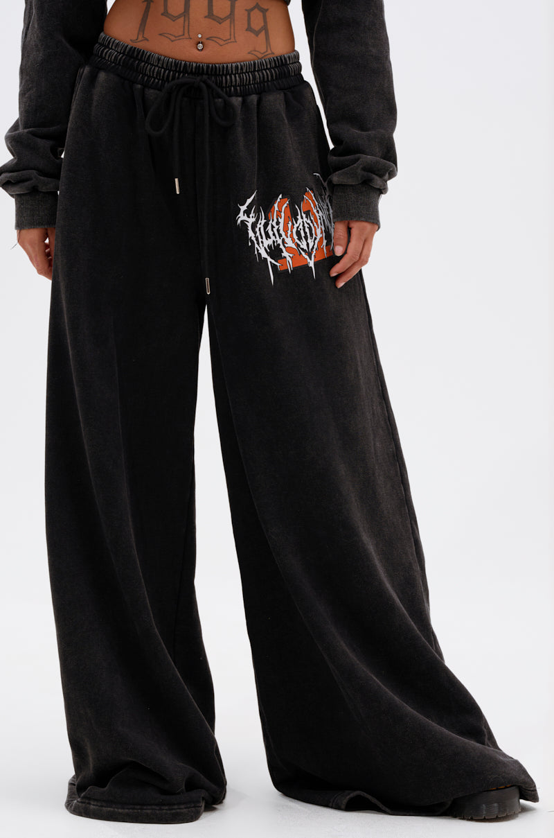 A person wears the LUCKY ELEVEN WIDE LEG JOGGER featuring a white and red graphic on the left thigh, paired with a matching black top. The cropped streetwear shot displays a "1999" tattoo on their lower torso. 2