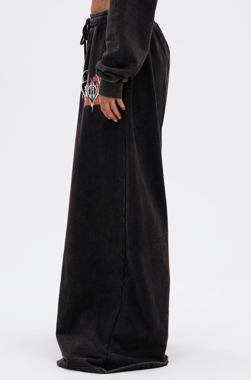 Someone wears the LUCKY ELEVEN WIDE LEG JOGGER—oversized black streetwear pants featuring a white and red graphic on the upper thigh. The wide-leg fit is so long, it covers the feet and reaches the ground. 4