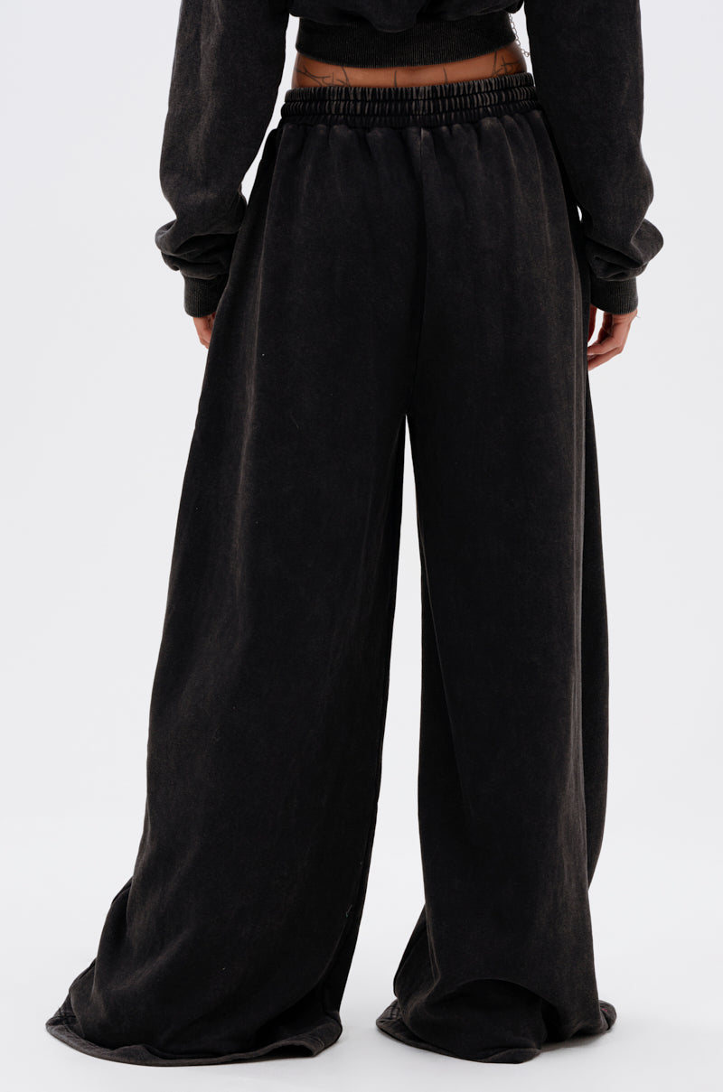 Viewed from the back, a person wears the LUCKY ELEVEN WIDE LEG JOGGER—loose-fitting, wide-leg pants with an elastic waistband that drape to the floor, partially covering their shoes. The plain light background adds a clean streetwear vibe. 5