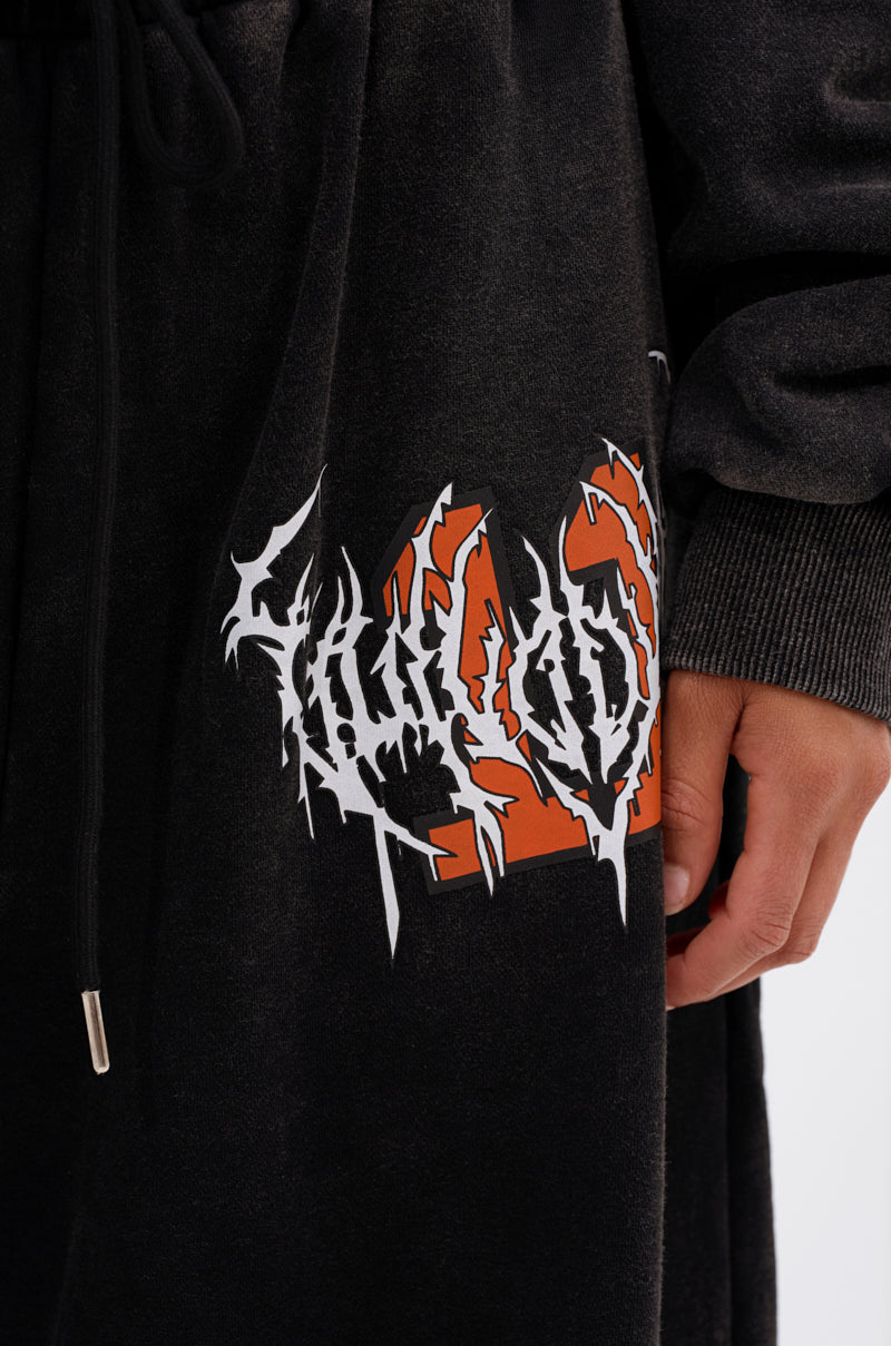 A close-up of a person’s hand resting on LUCKY ELEVEN WIDE LEG JOGGER pants in black acid-wash fleece, featuring a bold orange and white graphic with jagged lettering near the side pocket for a streetwear-inspired look. 7